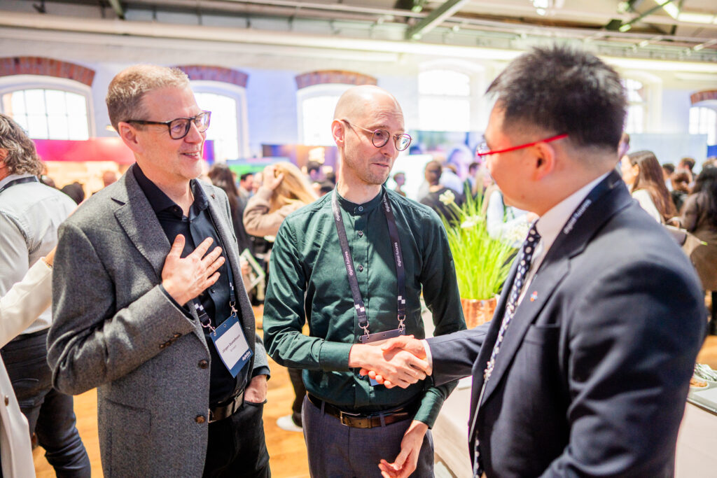 Three men in business attire interact at a networking event; two stand together while the third man, facing away from the camera, shakes hands with one of them. The background shows other attendees in a bright, open space.