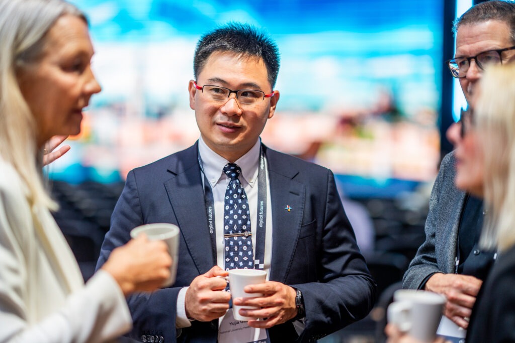 A man in a suit and tie holds a coffee cup whilst talking with three other people at a networking event. The group is indoors, and the background is blurred with blue and light tones.