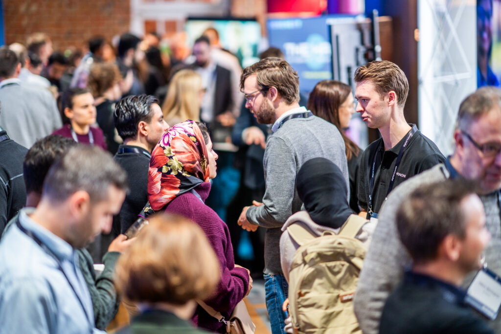 A large group of people mingle and converse at an indoor event or conference. Several attendees wear name badges and lanyards. The background is blurred, focusing on networking and conversation.