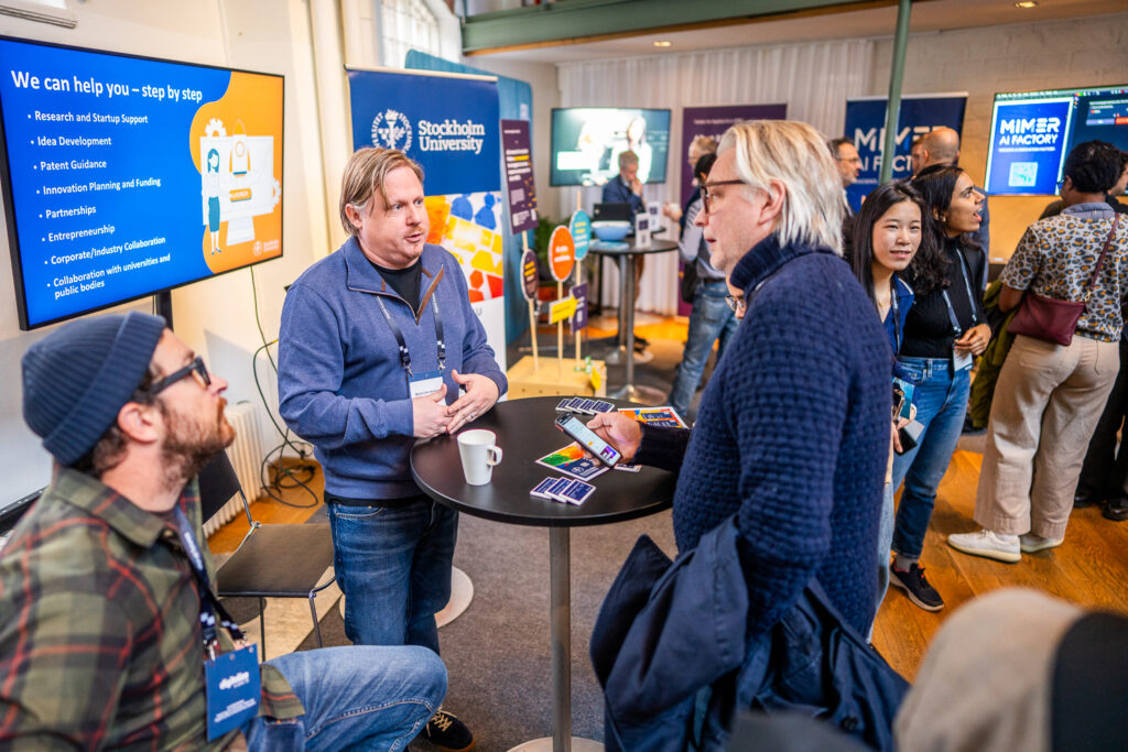 Two men converse at a round table during a busy expo. One holds cards, and a Stockholm University sign is visible in the background. Other people interact and visit stands in the brightly lit room.