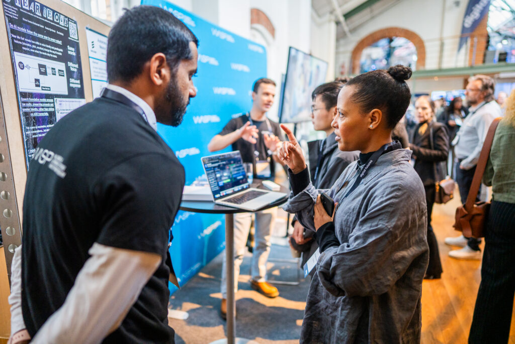 A woman speaks with a man in front of a digital display at a busy tech event. Other people and laptops are visible in the background, with a blue promotional banner behind them.