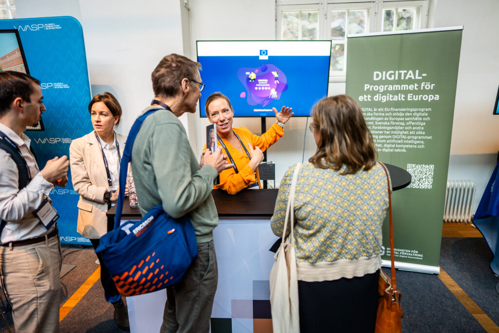 A woman in an orange shirt enthusiastically speaks to three people at a stand about a digital programme for Europe, with informational signs and a monitor displaying a digital map in the background.