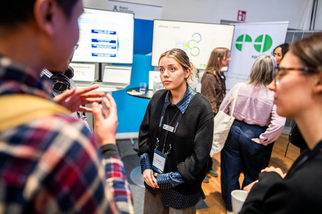 A woman with a conference badge listens attentively to a group at a networking event, with stands, posters, and other attendees in the background.