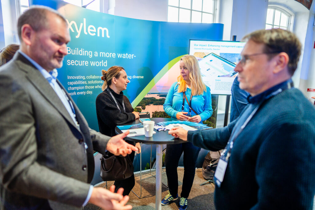 Four people converse at a stand with a blue “Xylem” banner about water solutions. Two men stand in the foreground, two women are at the display table with brochures and a cup, and a screen shows an informational graphic.