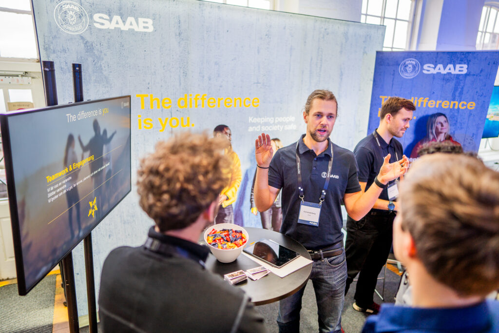 A man in a SAAB shirt speaks to two people at a stand with a display screen and a bowl of sweets. The stand features the SAAB logo and the slogan The difference is you. Other people interact nearby.