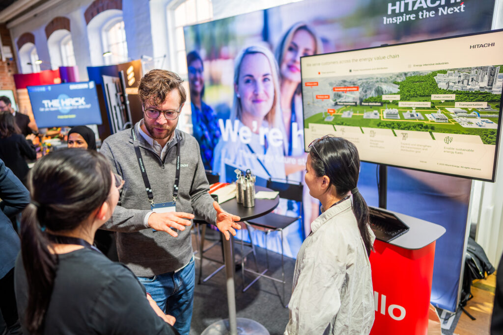 A man gestures while speaking to two women at a technology expo stand with a large screen displaying a presentation and a banner that reads We're here. Other attendees are visible in the background.