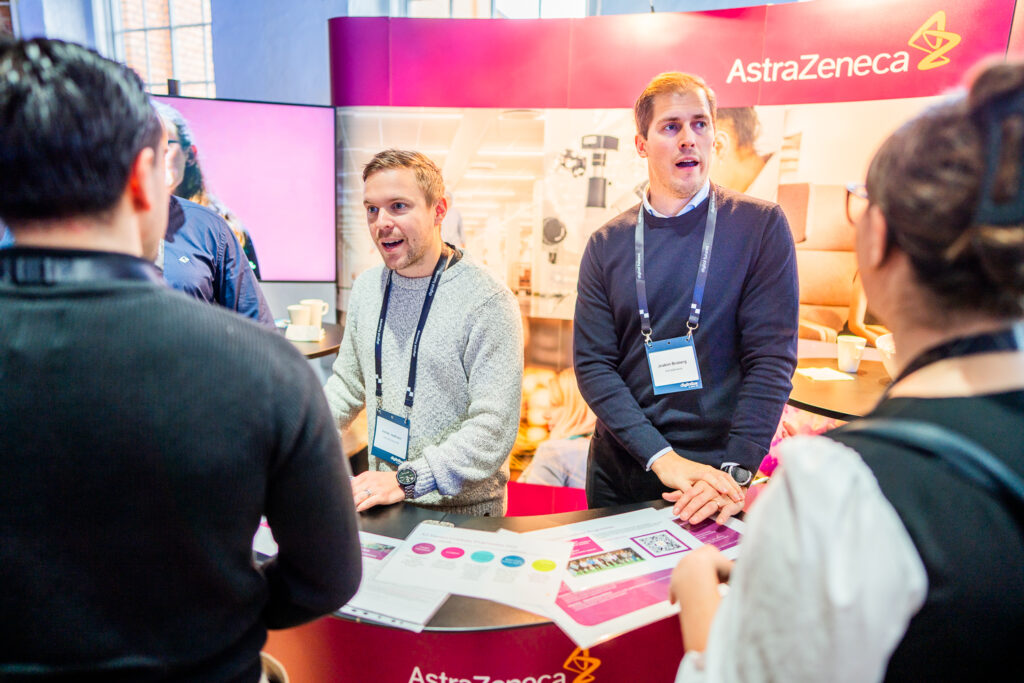 Two men stand behind an AstraZeneca stand, talking to attendees at an event. Promotional materials and informational brochures are displayed on the counter. People are engaged in conversation with the stand representatives.