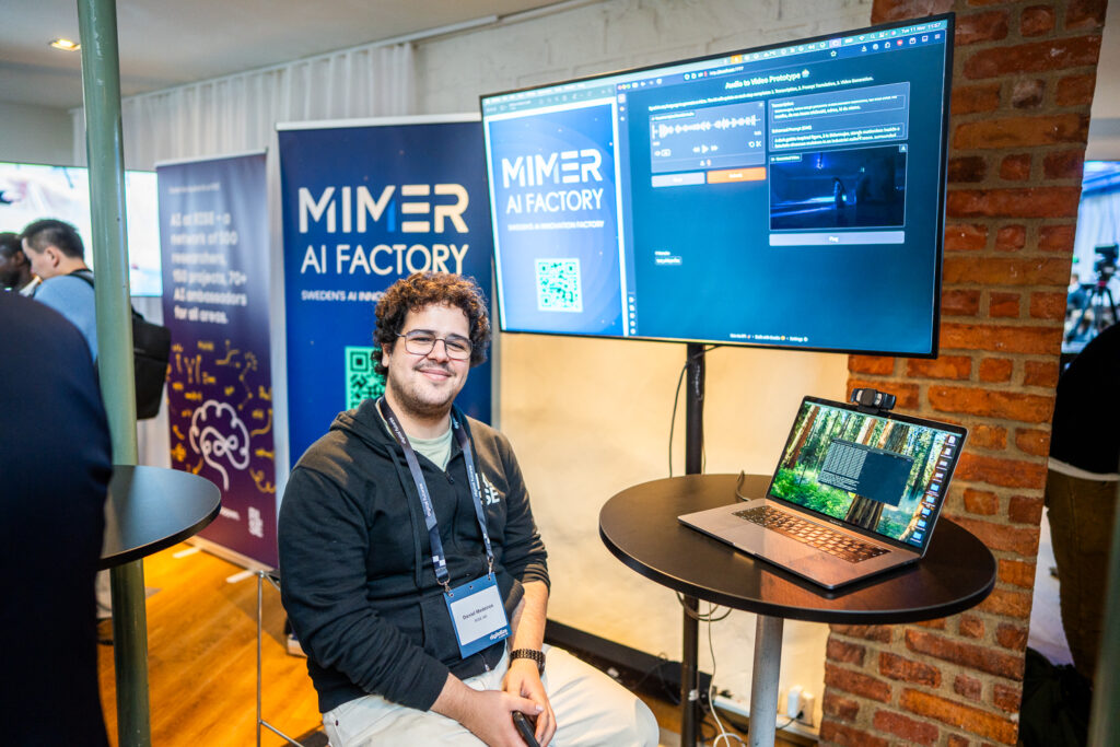 A smiling man with curly hair and glasses sits at a high table with a laptop, in front of MIMER AI FACTORY banners and screens showcasing software at a tech event.
