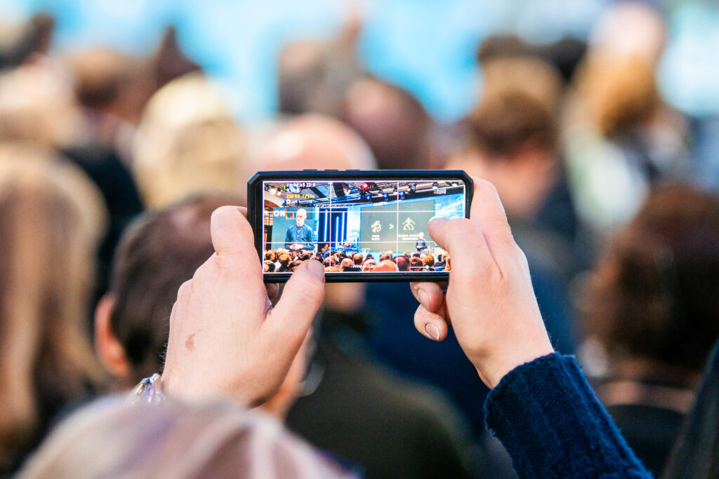 A person holds up a mobile phone to record or photograph a presentation or conference, with the stage and audience visible on the phone screen. The scene is filled with people in the background.