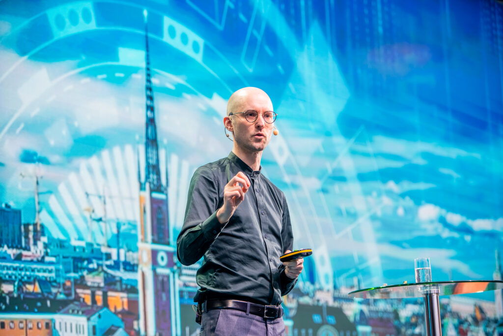 A bald man wearing glasses and a dark shirt speaks on stage, holding a mobile phone and gesturing with his hand. Behind him is a vibrant, futuristic cityscape backdrop with blue tones and digital graphics. A glass of water is on the table beside him.