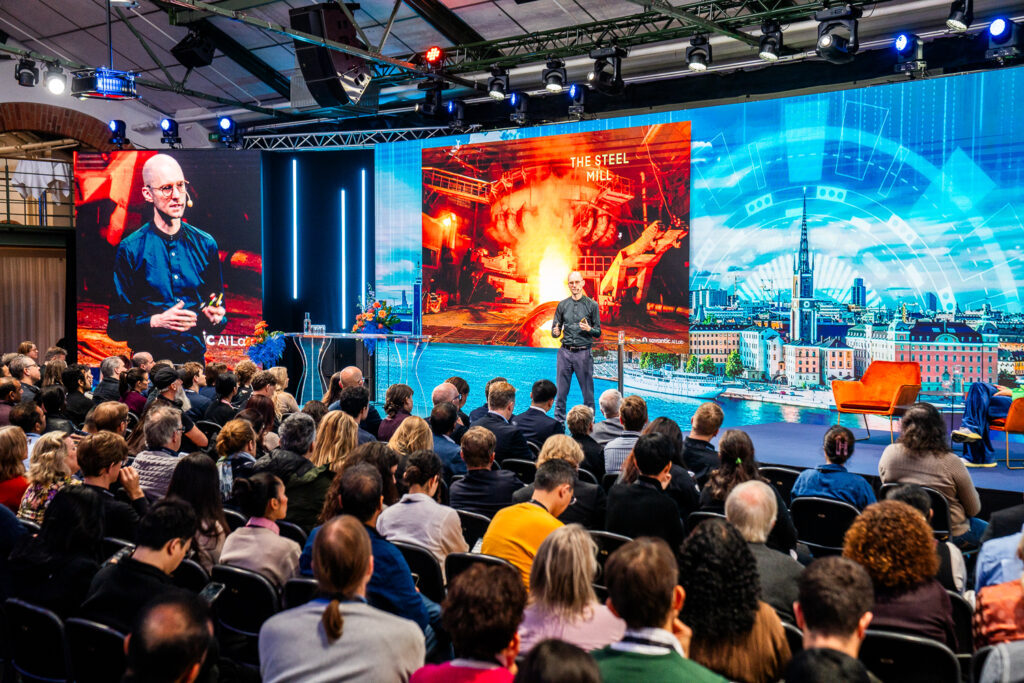 A speaker addresses a large seated audience at a conference. Behind the speaker, a screen displays a fiery steel mill image and the words THE STEEL NEED. The stage features modern décor and cityscape graphics.
