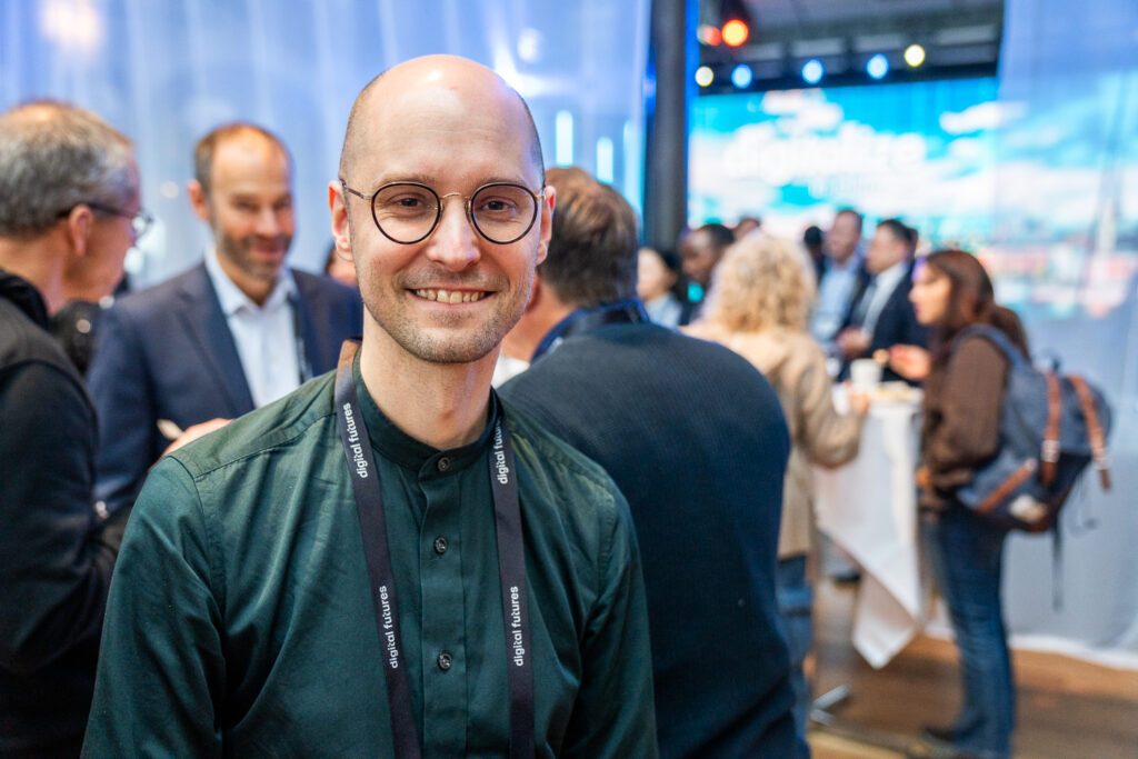 A smiling man with glasses stands in the foreground at a networking event, wearing a dark shirt and a lanyard. Several people are talking and mingling in the blurred background.
