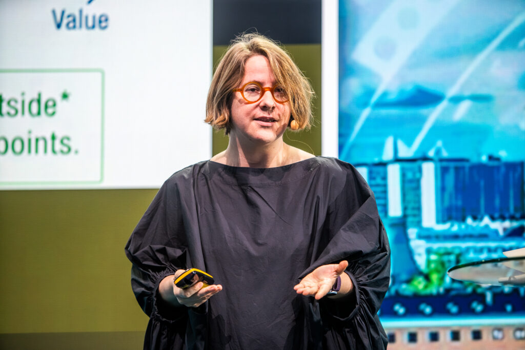 A person with short light brown hair and round glasses, wearing a loose black top, speaks on stage holding a device, with a screen displaying text and buildings in the background.