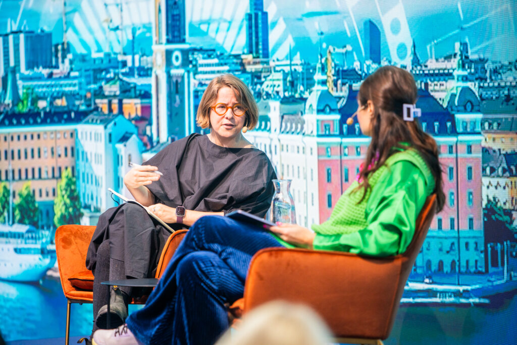 Two women sit in orange chairs and have a conversation on stage, with a colourful cityscape mural in the background. One woman gestures with her hand whilst the other listens attentively, holding a tablet.