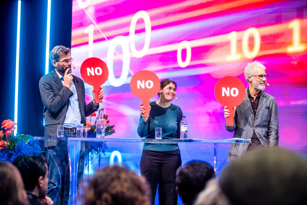 Three people stand behind a clear lectern, each holding up a red sign that says NO in white letters. They are on stage with a colourful digital background and an audience in front of them.