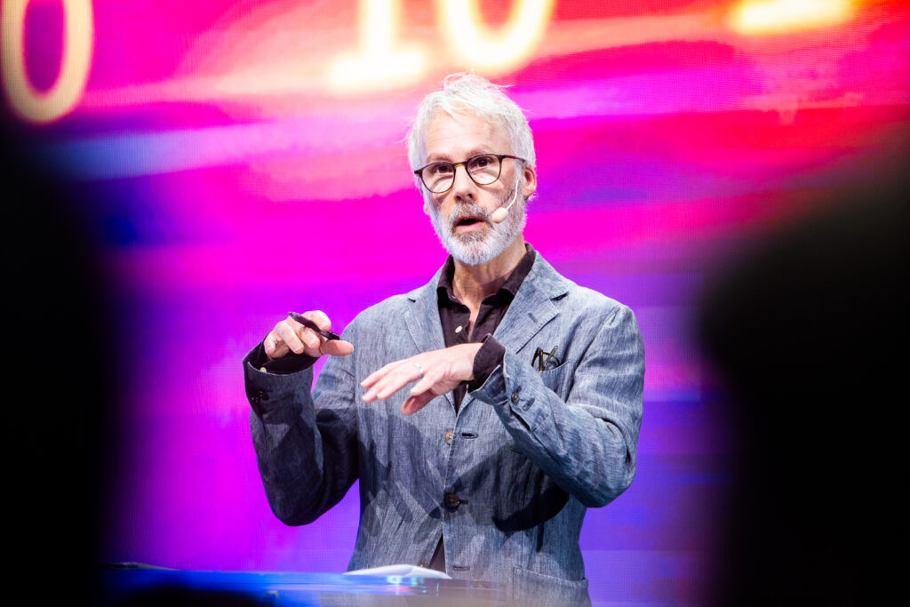 An older man with grey hair and beard, wearing glasses and a grey blazer, speaks on stage with a headset microphone against a vibrant pink and purple background.