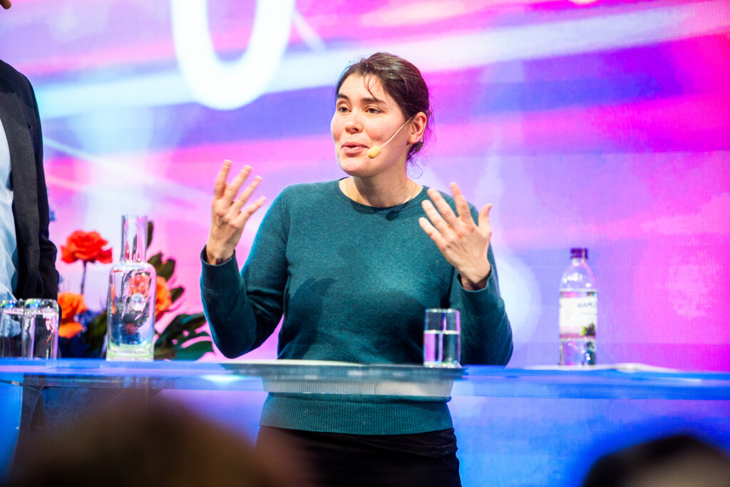 A woman wearing a green jumper speaks animatedly on stage with a microphone headset, gesturing with her hands. She stands behind a clear table with glasses of water, flowers, and a water bottle. The background is colourful and vibrant.