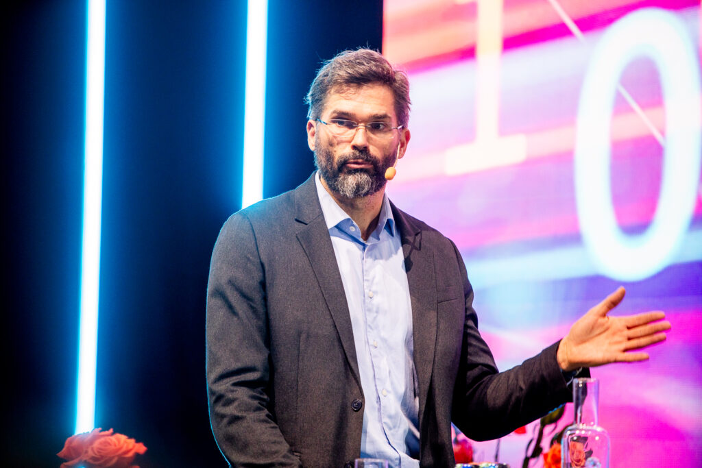 A man with glasses and a beard, wearing a suit jacket and a headset microphone, gestures whilst speaking on a brightly lit stage with colourful lights and abstract graphics in the background.