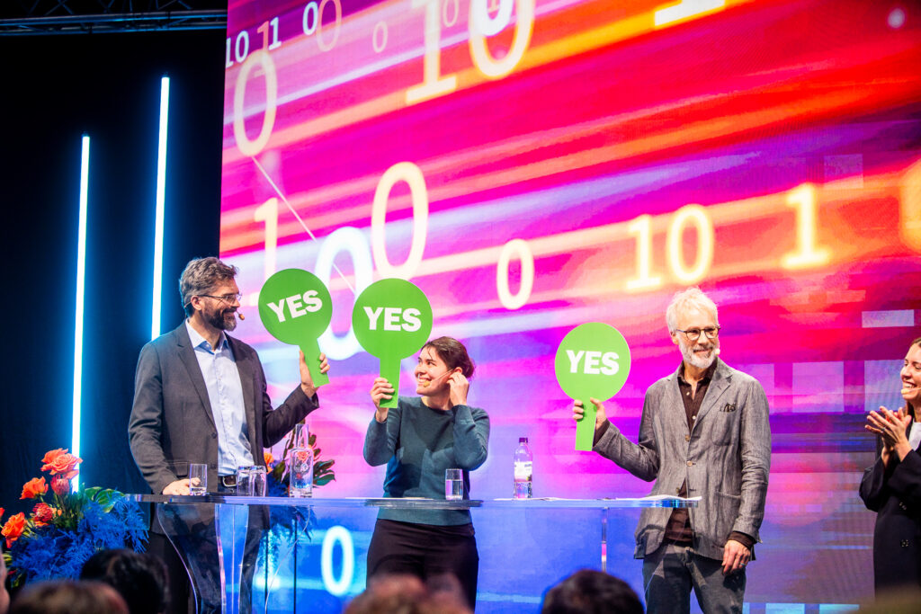 Three people stand behind a clear lectern on stage, each holding a green sign that says YES. A colourful background displays large numbers and bright lights. The audience is visible in the foreground.