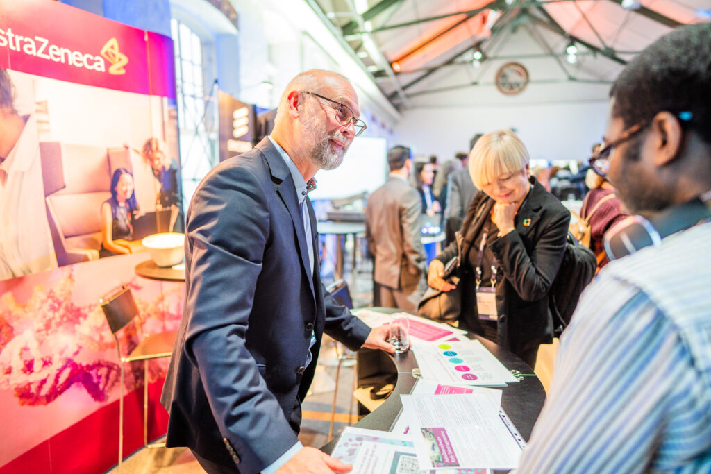 A man in a suit stands behind a stand with AstraZeneca branding, speaking to two people and handing out brochures at a busy indoor event or conference.