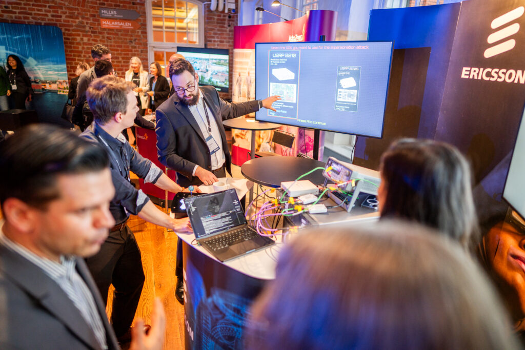 People at a tech expo stand interact with a display showing Ericsson equipment diagrams. One man gestures to the screen while others observe, and various devices and cables are arranged on the counter.