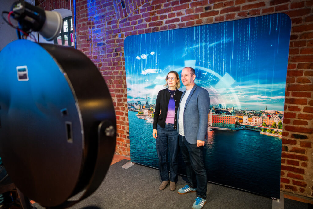 Two people stand side by side posing for a photo in front of a backdrop showing a cityscape with water and digital graphics, indoors with exposed brick walls and a camera set-up visible.