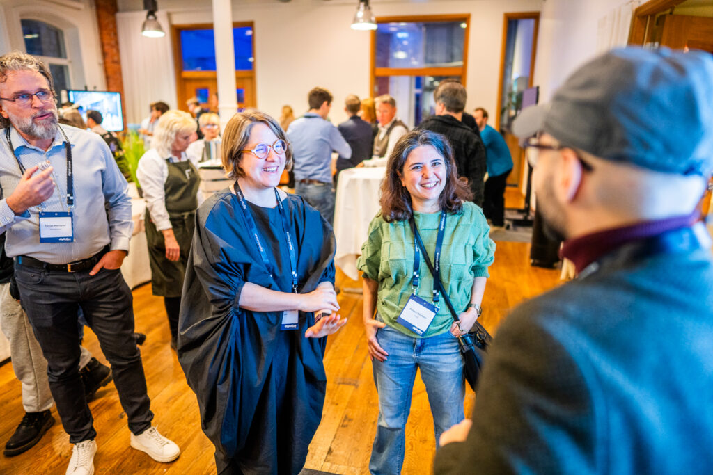 A group of people socialising at an indoor networking event; two women and a man are talking and smiling in the foreground, whilst others mingle in the background.
