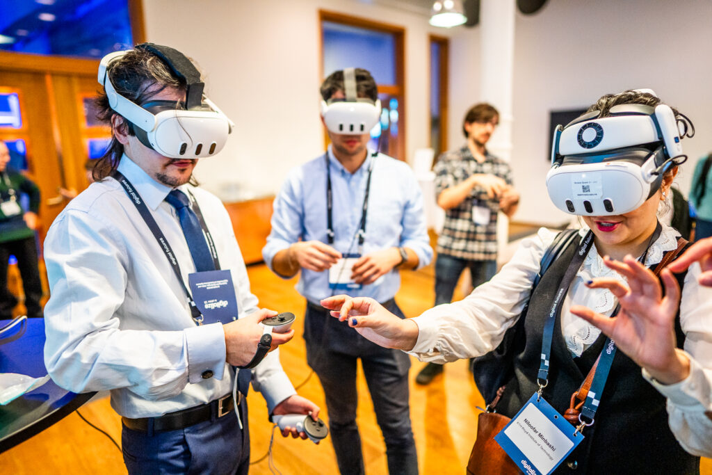 Three people in business attire wear virtual reality headsets and interact with controllers at an indoor event, whilst others watch in the background. The atmosphere appears lively and focused on technology.