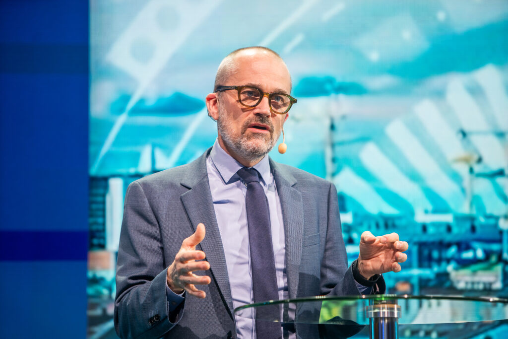A man in a suit and glasses speaks on stage with a headset microphone, gesturing with his hands. A colourful, blurred cityscape and abstract graphics are visible in the background.