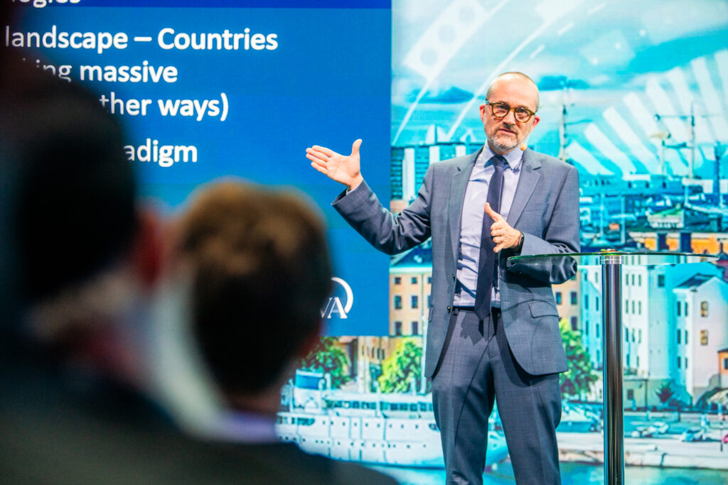 A man in a suit and glasses gestures whilst speaking on stage in front of a blue presentation screen and cityscape backdrop, with audience members visible in the foreground.