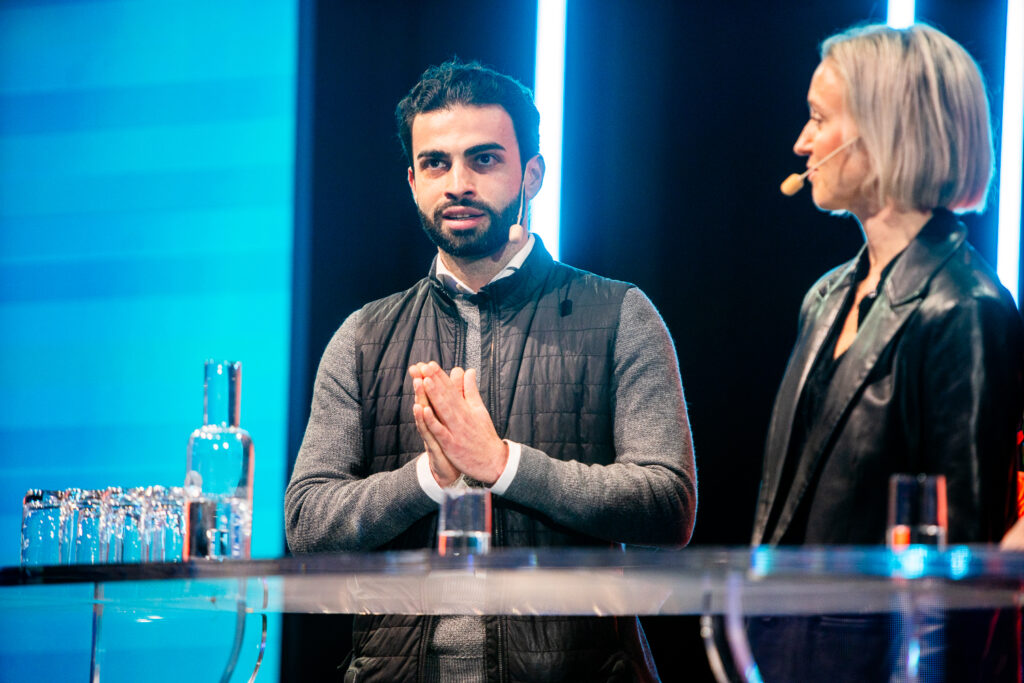 Two people stand behind a clear table with glasses and a water carafe, speaking into headset microphones on a brightly lit stage with blue vertical lights in the background.