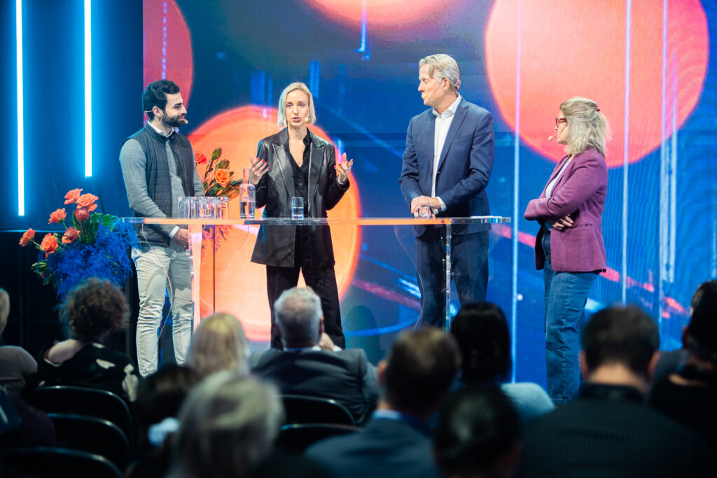 Four people stand around a clear table on stage, engaged in discussion, with a colourful blue and orange backdrop and an audience seated in front of them. Flowers are arranged to the side of the table.