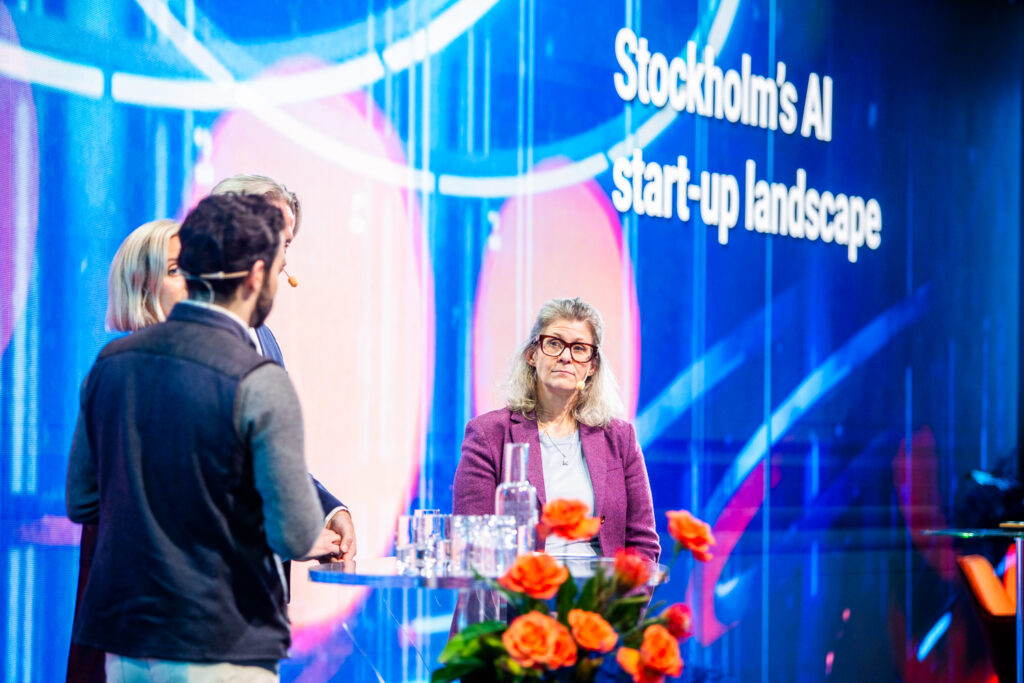 Four people stand onstage having a discussion at a tech event. The background screen reads Stockholm’s AI Start-up landscape. A table with water glasses and a vase of orange flowers is in the foreground.