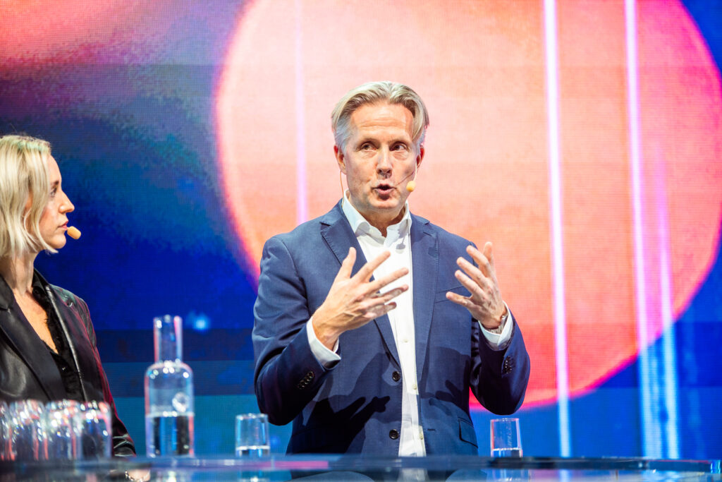 A man in a blue suit gestures whilst speaking on stage with a woman beside him; both wear headset microphones, and colourful lights create a vibrant background.