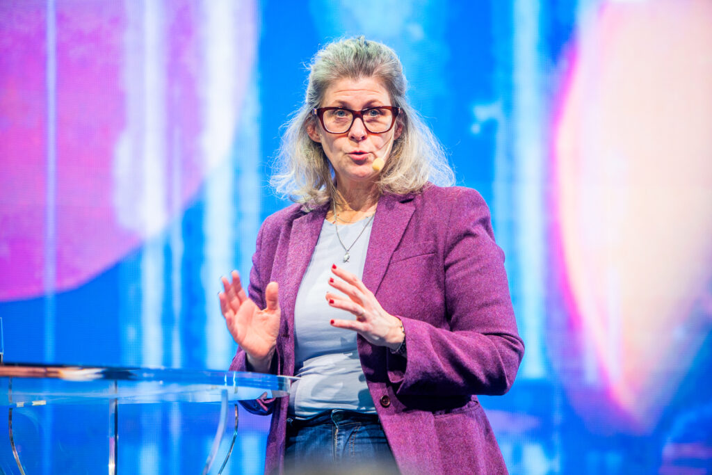 A woman with grey hair and glasses, wearing a purple blazer and light shirt, speaks on stage with a colourful, blurry blue and pink background. She gestures with her hands near a clear lectern.