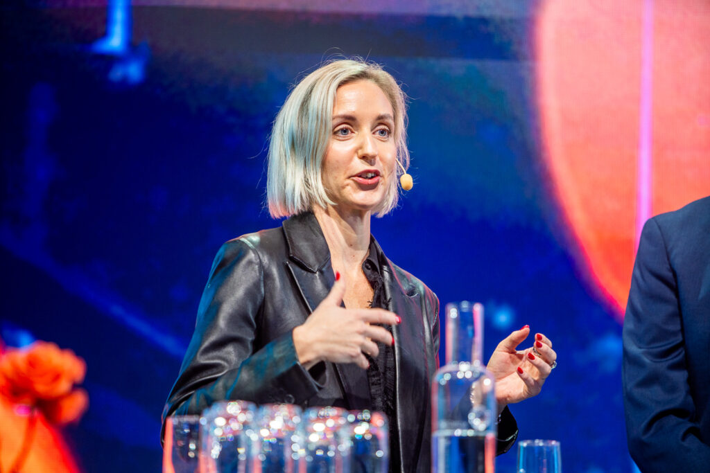 A woman with short blonde hair speaks on stage wearing a dark jacket and a headset microphone. She gestures with her hands, and glass bottles are on the table in front of her. The background is colourful and abstract.