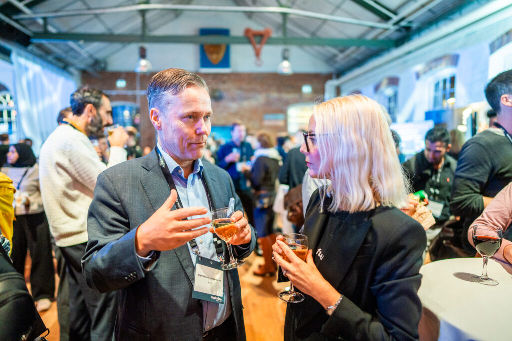 Two people in business attire stand in a brightly lit event space, holding drinks and engaged in conversation, with other attendees and tables visible in the background.