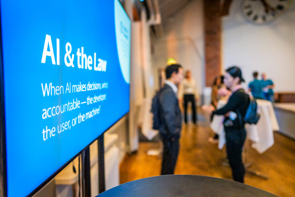 A screen displays AI & the Law as people converse in a bright room with wooden floors and a large clock on the wall in the background. The event discusses AI decision-making and accountability.