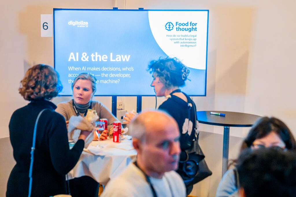 Four people sit and talk at a round table in front of a large screen displaying a discussion topic titled AI & the Law at an event. The screen asks about legal accountability for AI decisions.