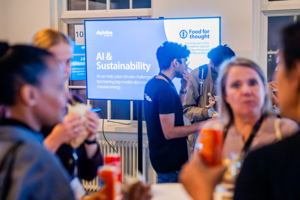 A group of people socialises at an event in front of a screen displaying “AI & Sustainability” and “Food for thought,” discussing the impact of AI on climate challenges and energy consumption.