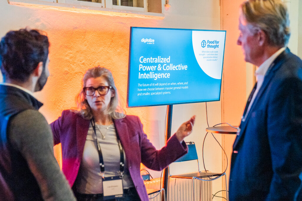 Three people have a discussion in front of a screen displaying a presentation titled Centralised Power & Collective Intelligence at an indoor event. The woman in the centre gestures with her hands whilst talking.