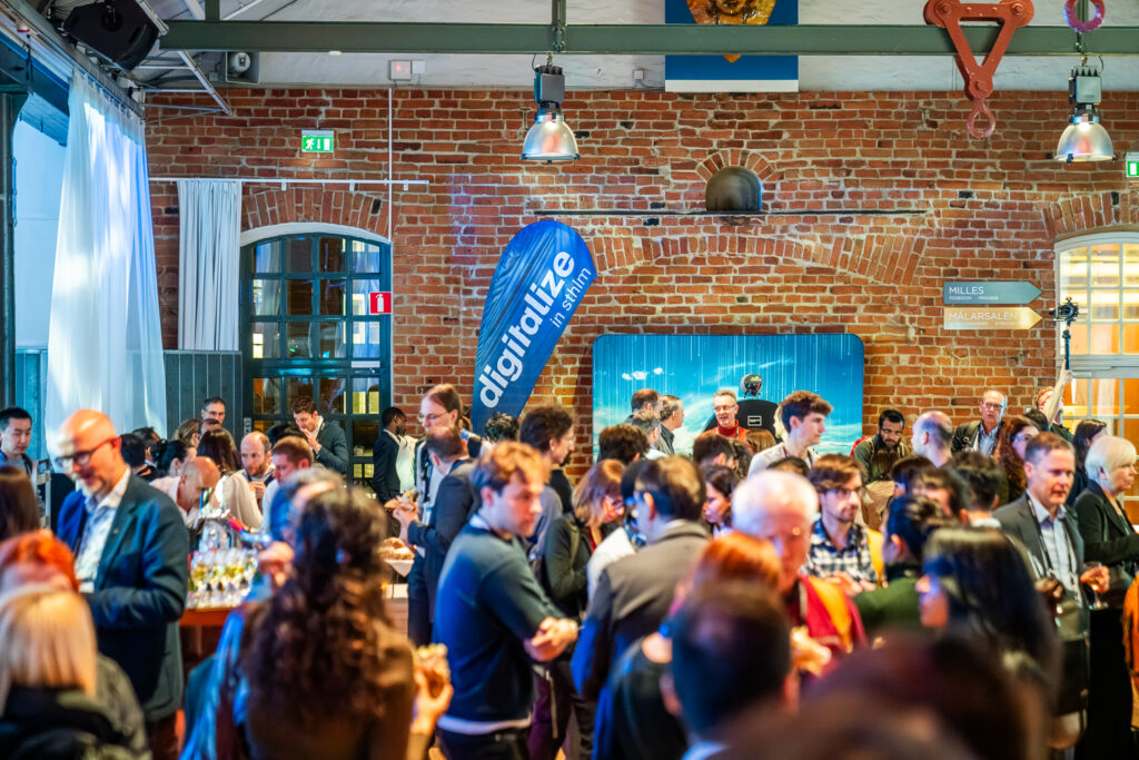A large group of people socialise at an indoor event with exposed brick walls. A blue digitalise banner is visible, and guests are dressed in business attire. The atmosphere is lively and professional.