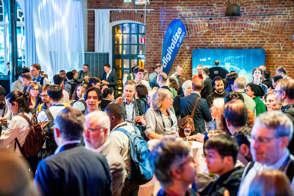 A large group of people socialising at an indoor networking event, with some holding drinks. There is a “digitalise” banner and brick walls in the background, along with white curtains and a modern, tech-themed atmosphere.
