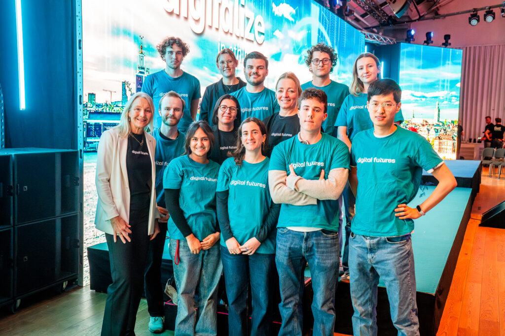 A group of people wearing teal digital futures shirts pose and smile on a stage, alongside a woman in a white blazer, with a bright digital-themed background behind them.