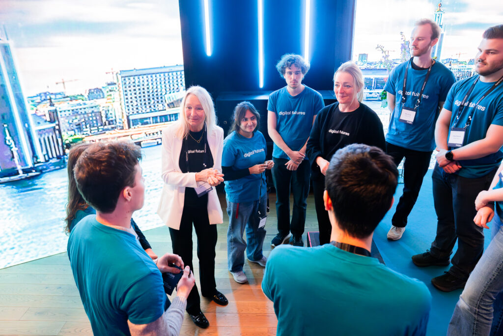 A group of people wearing blue digital talent shirts and name tags stand in a circle on a stage, talking and smiling, with a cityscape background and blue vertical lights behind them.