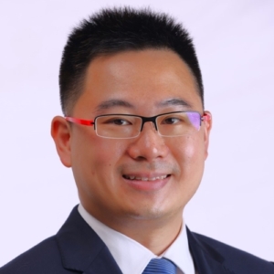 A man with short black hair and glasses, wearing a dark suit, white shirt, and blue tie, smiling in front of a plain light background.