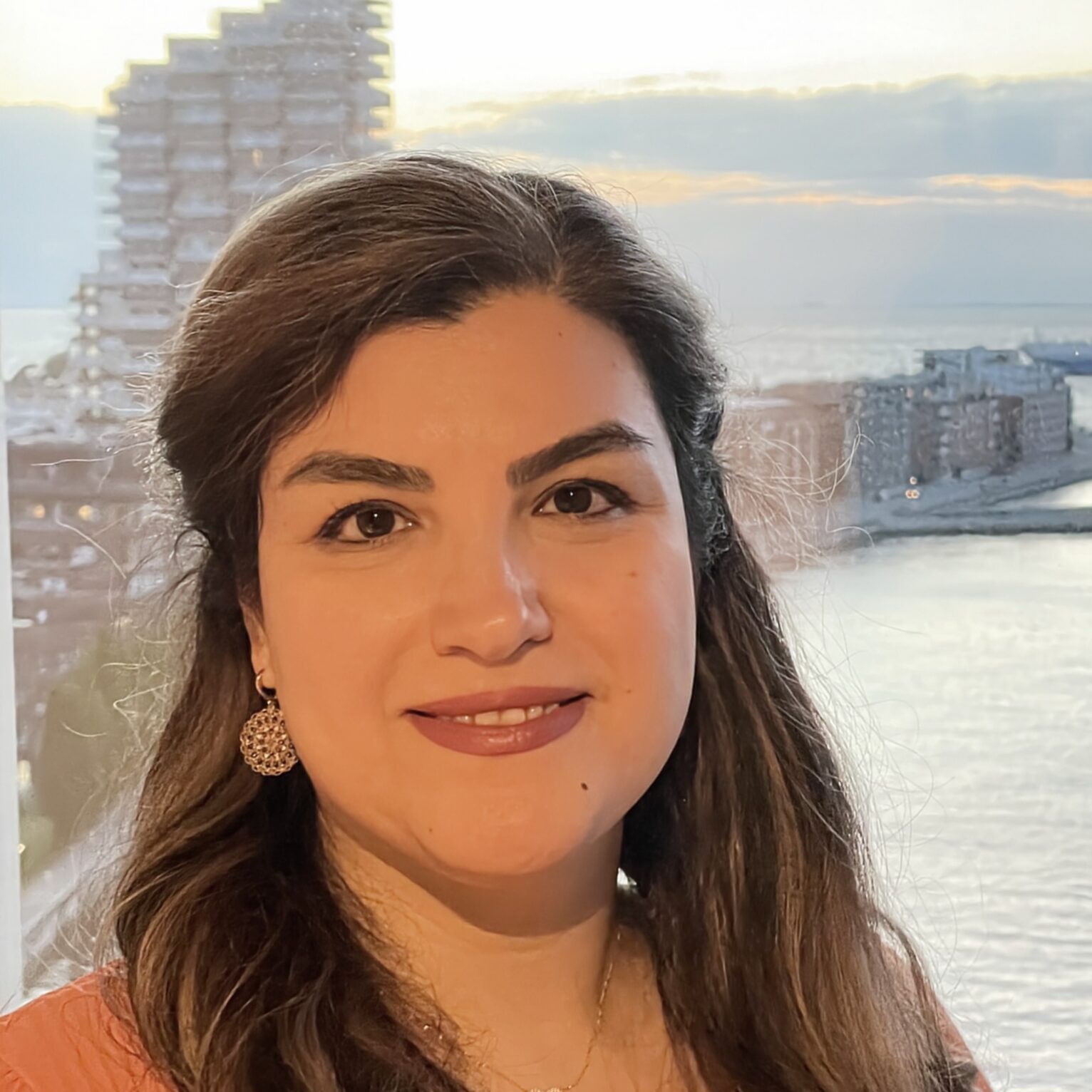 A woman with long brown hair and a coral top smiles at the camera, standing in front of a window overlooking a city waterfront at sunset, with buildings and a ship visible in the background.