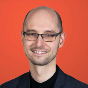 A smiling man with glasses, a bald head, and light facial hair is wearing a dark blazer and black shirt, standing in front of an orange background.