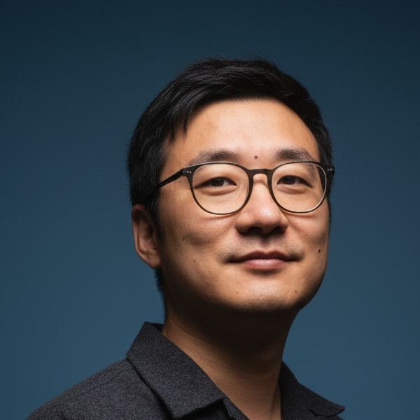 A man with short black hair and glasses wearing a dark button-up shirt poses confidently against a plain blue background.