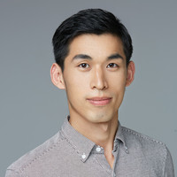 A young man with short black hair, wearing a light grey button-up shirt, poses in front of a plain grey background, looking directly at the camera with a neutral expression.